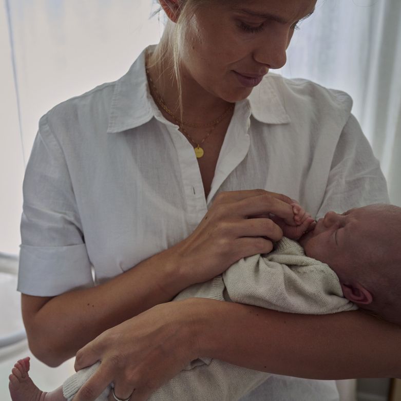 A woman in a white shirt gently cradles a sleeping baby, gazing at the infant with a nurturing expression.
