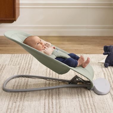 Baby resting in a green baby bouncer on a beige textured rug. The baby is looking to the side with a relaxed expression.