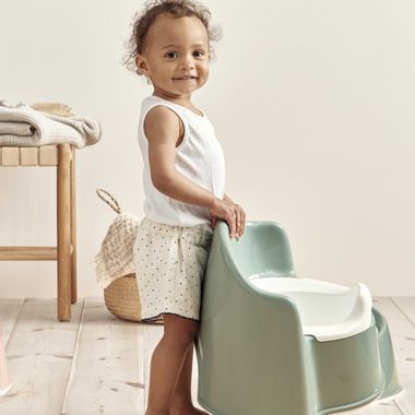 A toddler stands smiling next to a green potty chair in a bright room with wooden floors and a wicker basket in the background.
