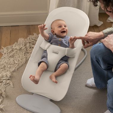 A baby smiling in a white bouncer reaches for a person's finger, on a wooden floor with a cozy rug nearby.