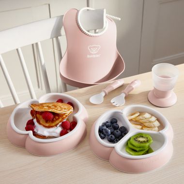 Pink baby feeding set with two dishes: pancakes with raspberries, blueberries with bananas and kiwi. Includes a bib, cup, spoon, and fork.