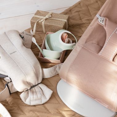 Baby essentials arranged on a quilt, including a pink baby bouncer, feeding set, swaddle, and a wrapped gift box with a beige ribbon.