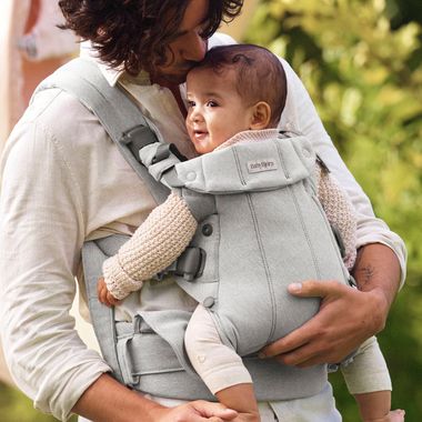 Parent holding a smiling baby in a grey carrier, surrounded by greenery, both wearing soft, light-colored clothes.