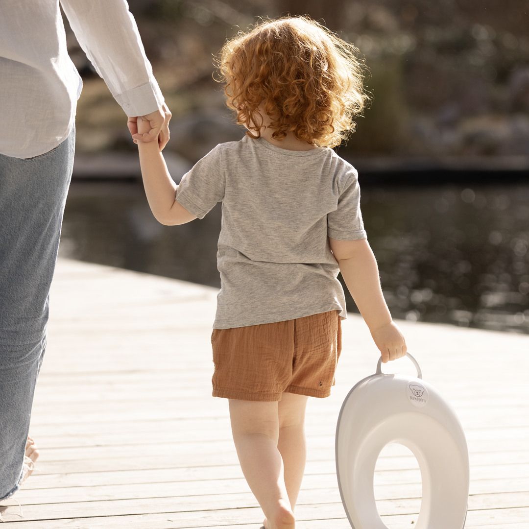 Child with red curly hair holding an adult's hand, carrying a potty seat, walking on a wooden deck near a body of water.