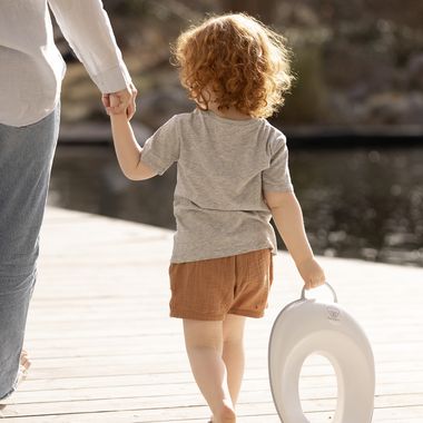 Child with red curly hair holding an adult's hand, carrying a potty seat, walking on a wooden deck near a body of water.