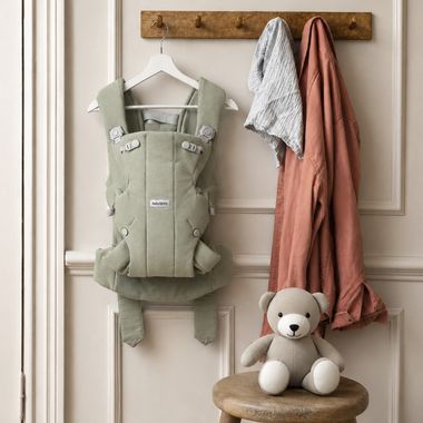 A green baby carrier hangs on a wooden hook, next to draped clothes, above a stool with a stuffed teddy bear on it.