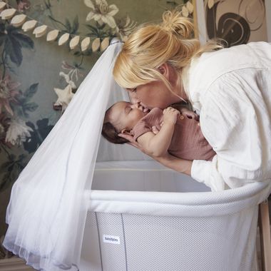 A woman gently kisses a baby's nose in a white bassinet with a canopy. The room has floral wallpaper and garland decorations.