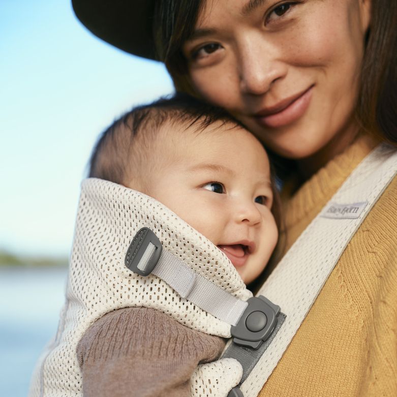 A woman smiles while holding a baby in a carrier outdoors. Both look content and happy. The background shows a blurred natural setting.