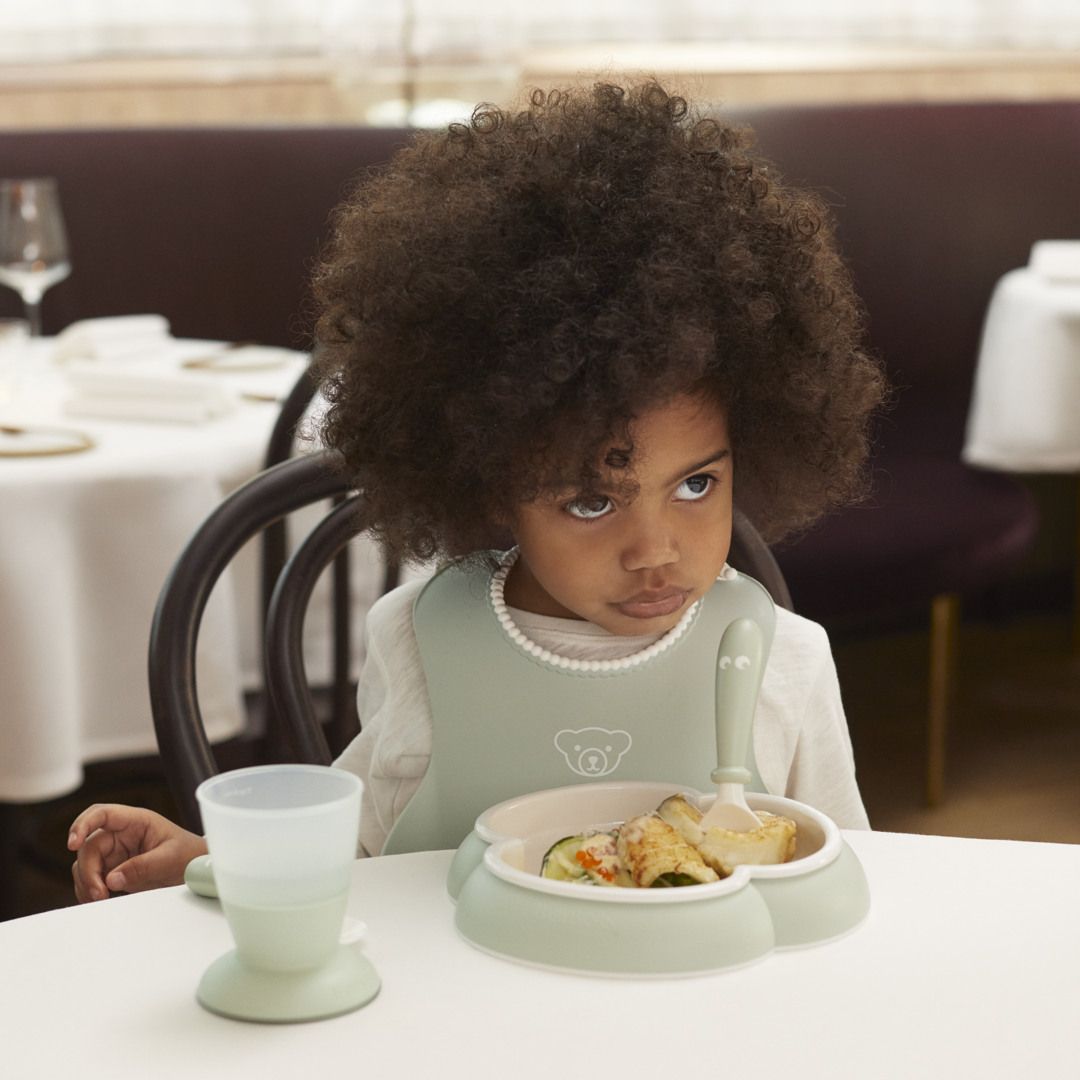 A child with curly hair sits at a table in a restaurant, wearing a bib and looking unimpressed, with a plate of food and a cup in front of them.