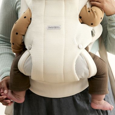 A baby in a beige carrier, wearing brown and tan clothes with black polka dots, being held by an adult in a light green sweater.