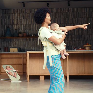 A person wearing colorful pants holds a baby in a carrier while pointing, with another baby in a bouncer in the background in a modern kitchen.
