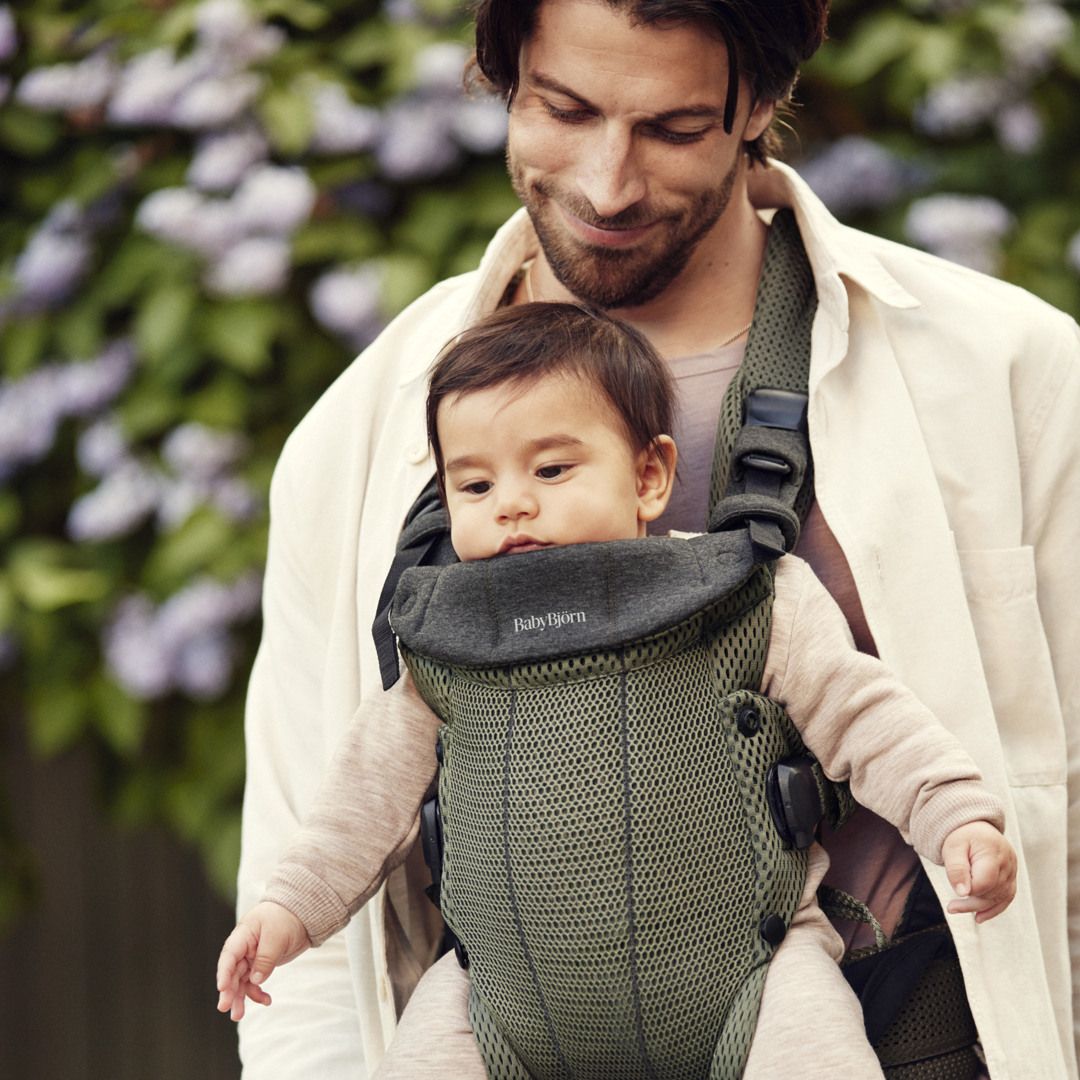 Man smiling at a baby in a green mesh BabyBjörn carrier outdoors, with purple flowers in the background.