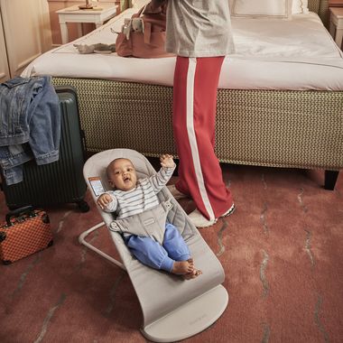 Baby in a bouncer on a carpeted floor, near a person packing a suitcase on a bed. Clothing and luggage are visible beside them.