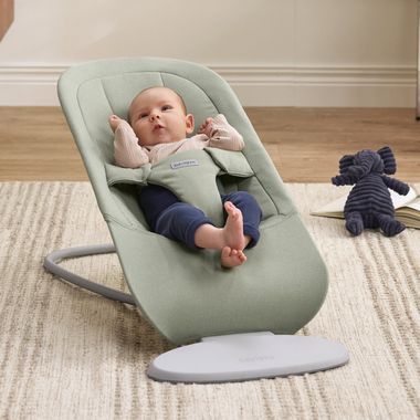 A baby in a light green bouncer, wearing a pink top and blue pants, is lying on a beige carpet with a stuffed elephant toy nearby.