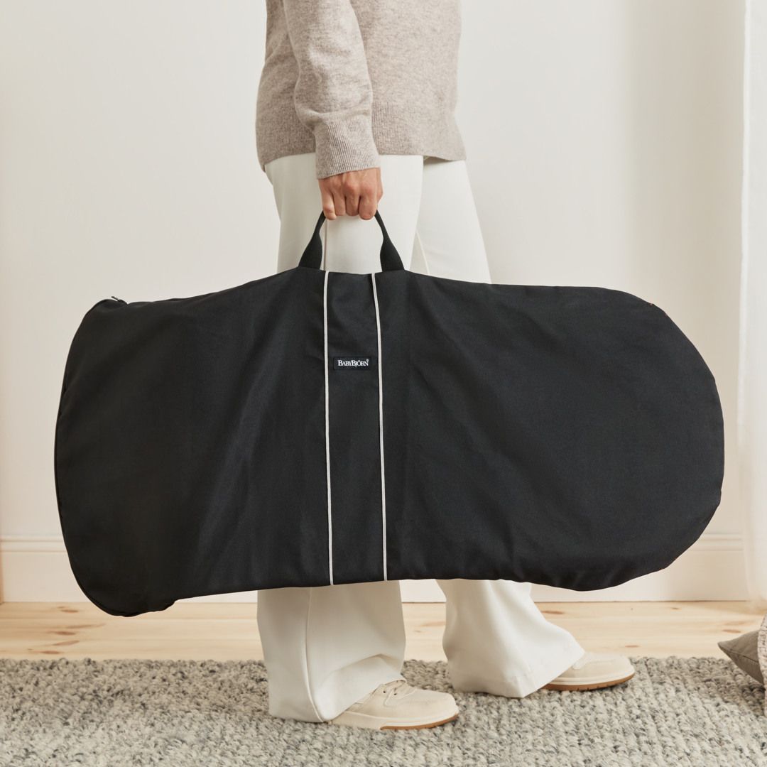Person holding a large black carrying bag for bouncer with handles, standing on a wooden floor in a light-colored room.