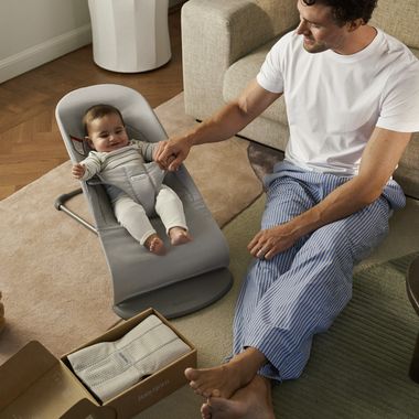 A man in pajamas holds the hand of a smiling baby in a gray bouncer. A cardboard box with a blanket is on the floor nearby.