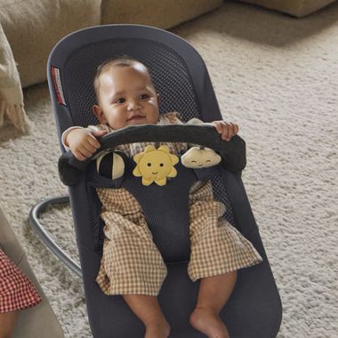 A baby sitting in a bouncer chair, wearing a plaid outfit and holding onto a toy bar with attached plush sun and cloud.