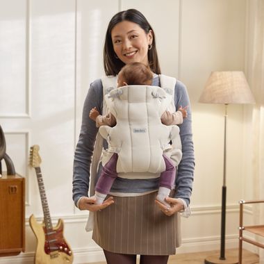 A woman smiles while carrying a baby in a white carrier in a cozy room with a guitar and floor lamp in the background.