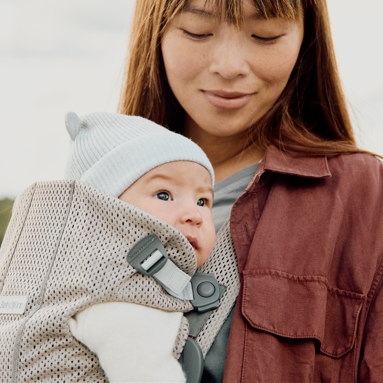 A woman lovingly gazes at a baby wearing a light blue beanie and strapped into a cozy, gray baby carrier.