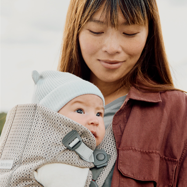 A woman gently holds a baby wearing a light blue hat in a grey baby carrier. The woman is smiling softly, looking down at the baby.