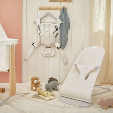 Cozy nursery with a beige baby bouncer, a hanging baby carrier, soft toys, and books on a plush rug. Neutral tones create a warm atmosphere.