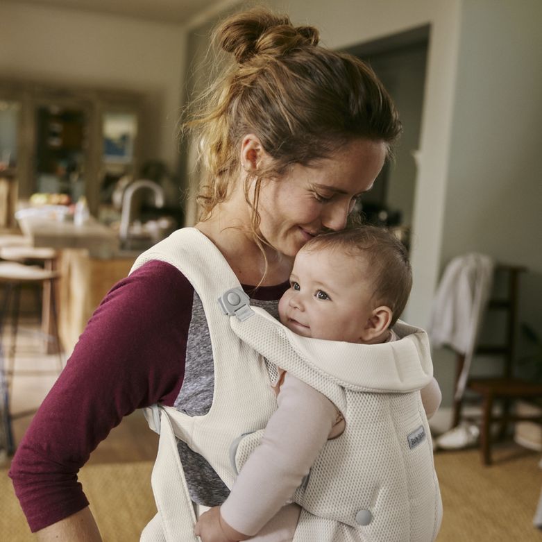 A woman smiles and gently embraces a baby in a carrier inside a cozy home setting.