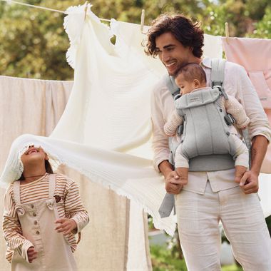 Man holding baby in a gray carrier, smiling at a child playing with a sheet on a clothesline in a sunny garden.