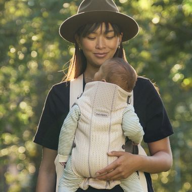 A woman outdoors, wearing a hat, holds a baby in a striped outfit secured in a baby carrier, surrounded by dappled sunlight and greenery.