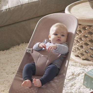 A baby in a pink mesh bouncer on a soft carpet, gazing upwards, with a round wooden table in the background.