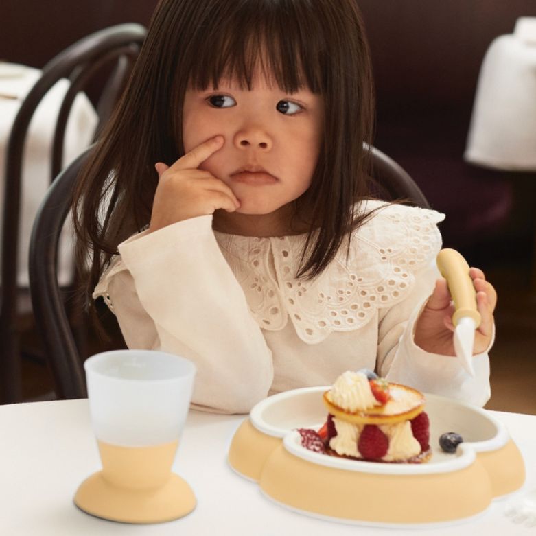 A young girl with long hair sits at a table with a layered dessert and a cup, looking thoughtful, holding a fork in one hand.