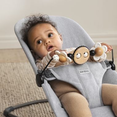Baby in a gray bouncer with a wooden toy bar, looking up. The baby has curly hair and wears a striped shirt and brown pants, seated on a carpet.