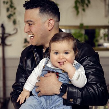 Smiling man holds a baby dressed in denim overalls, with greenery in the background.
