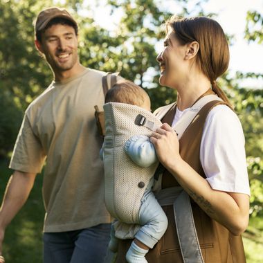 Smiling parents enjoy a sunny day in the park, carrying a baby in a cream baby carrier mini.