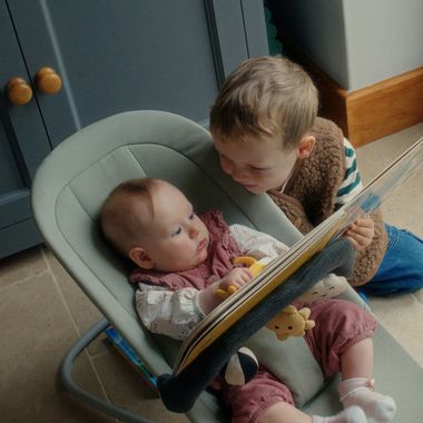 A young child reads a book to a baby sitting in a bouncer, with soft toys attached, in a cozy indoor setting.