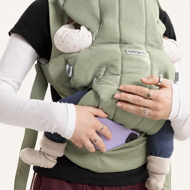 A person wearing a green baby carrier holding a baby, with their hand on a purple phone. The baby is dressed in polka-dot and plain clothes.