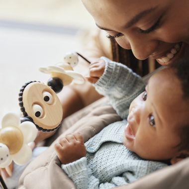 Smiling baby in a blue sweater playing with a wooden toy, closely watched by an adult.