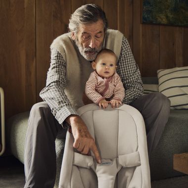 Grandfather with gray hair sits on a couch, holding a baby in a striped outfit who is standing on a fabric chair indoors.
