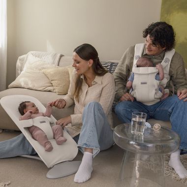 A couple sits on a couch with a baby. The woman smiles at a baby in a bouncer, while the man holds another baby in a baby carrier.