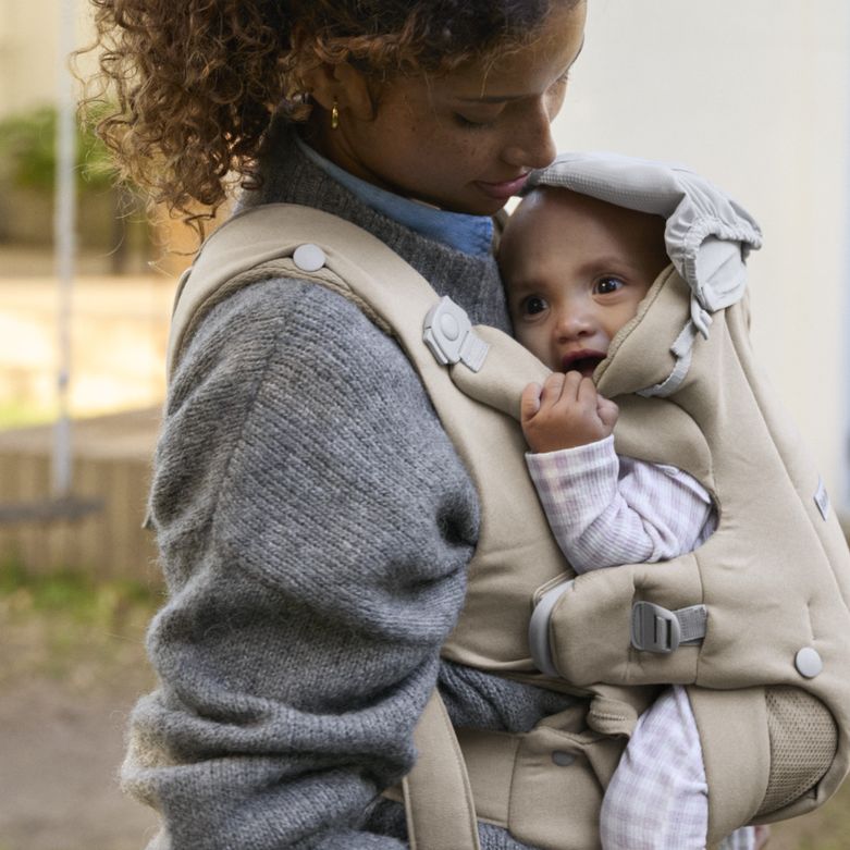 A woman wearing a grey sweater holds a baby in a beige carrier. The baby, dressed in pink, looks curiously out from the carrier's hood.