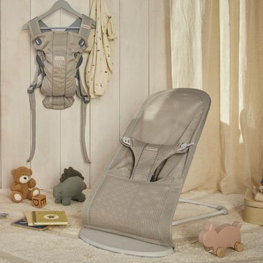 Beige baby bouncer and carrier set in a cozy nursery with toys and books on a soft carpet, next to light curtains.