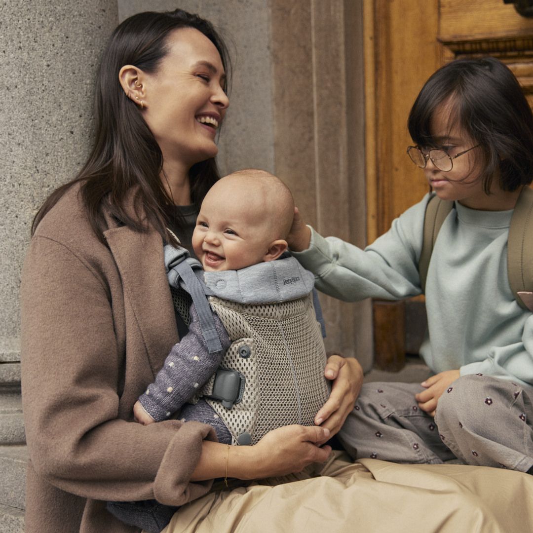 A smiling woman holds a baby in a carrier, while a child with glasses gently touches the baby's head. They are sitting near a stone column.