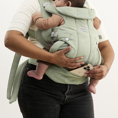 An adult carrying a baby in a mint green baby carrier, holding a smartphone in one hand. The baby is wearing brown clothes.