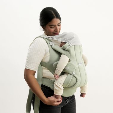 A woman in a white shirt holds a baby in a green carrier with a sunshade, gazing affectionately at the sleeping infant.