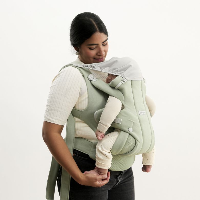 A woman in a white shirt holds a baby in a green carrier with a sunshade, gazing affectionately at the sleeping infant.