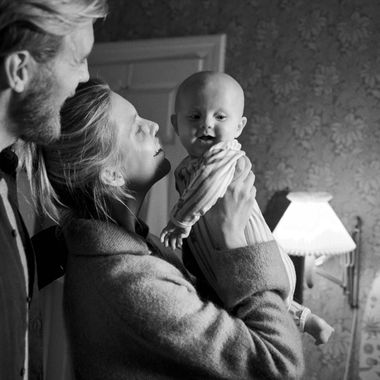 A woman holds a smiling baby in her arms while a man stands beside them in a warmly lit room with floral wallpaper and a lamp.