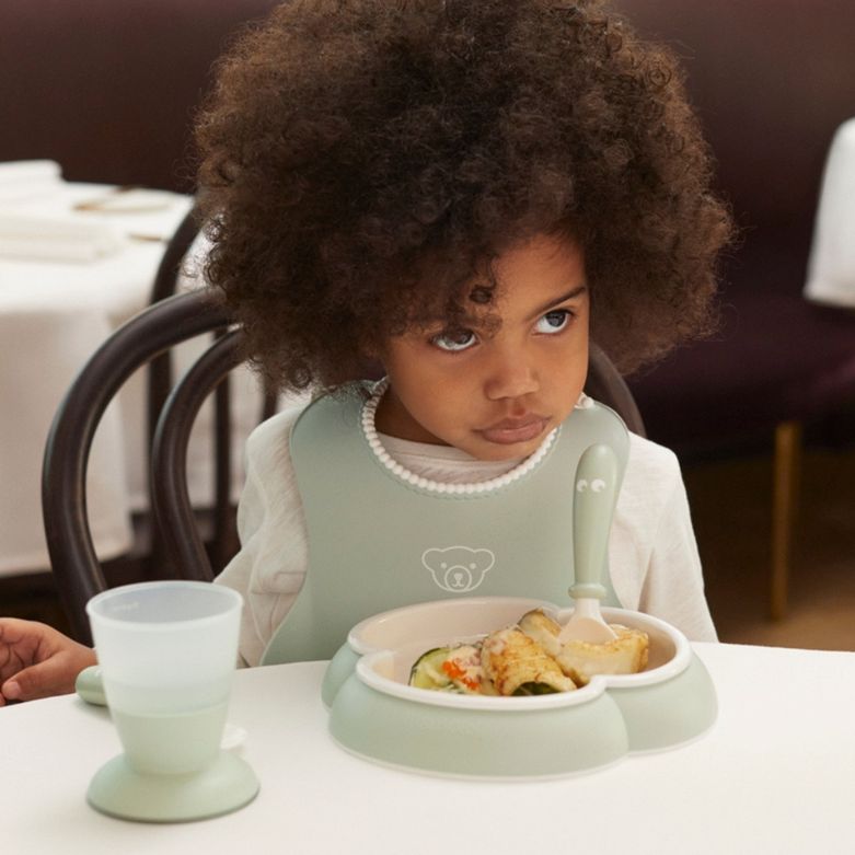 A child with curly hair sits at a table, wearing a bib, looking upward with a serious expression, while holding a fork over a partially eaten meal.