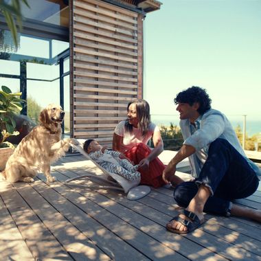 A couple sits on a wooden deck with a baby in a bouncer, interacting with a friendly golden retriever, surrounded by greenery and a modern house.