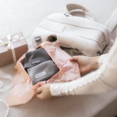 Hands unwrapping baby hats in pink tissue paper, with a gift box and diaper bag on a table.