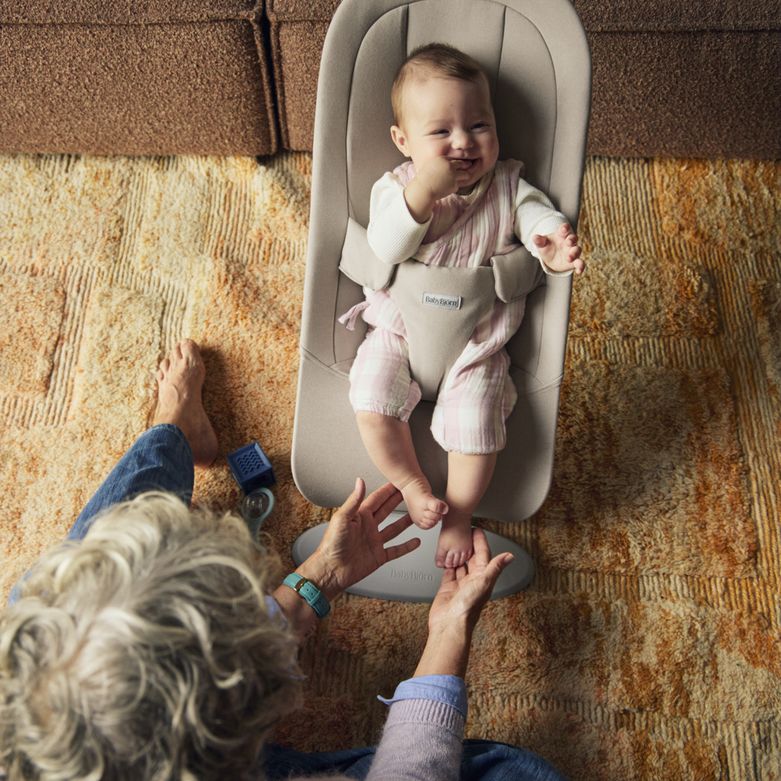 A baby in pink overalls sits smiling in a beige baby chair, while an adult reaches out towards them on a patterned orange carpet.