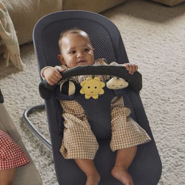 Baby in a checkered outfit sits in a bouncer holding a toy bar with plush sun and cloud, on a light carpeted floor.